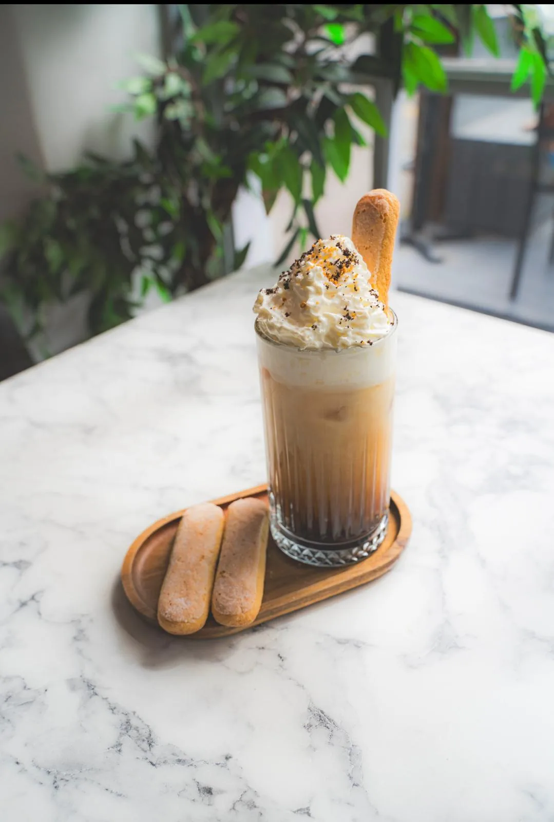 Iced coffee at TenTen in a fluted glass, topped with cream and cocoa, served with savoiardi biscuits on a wooden tray.