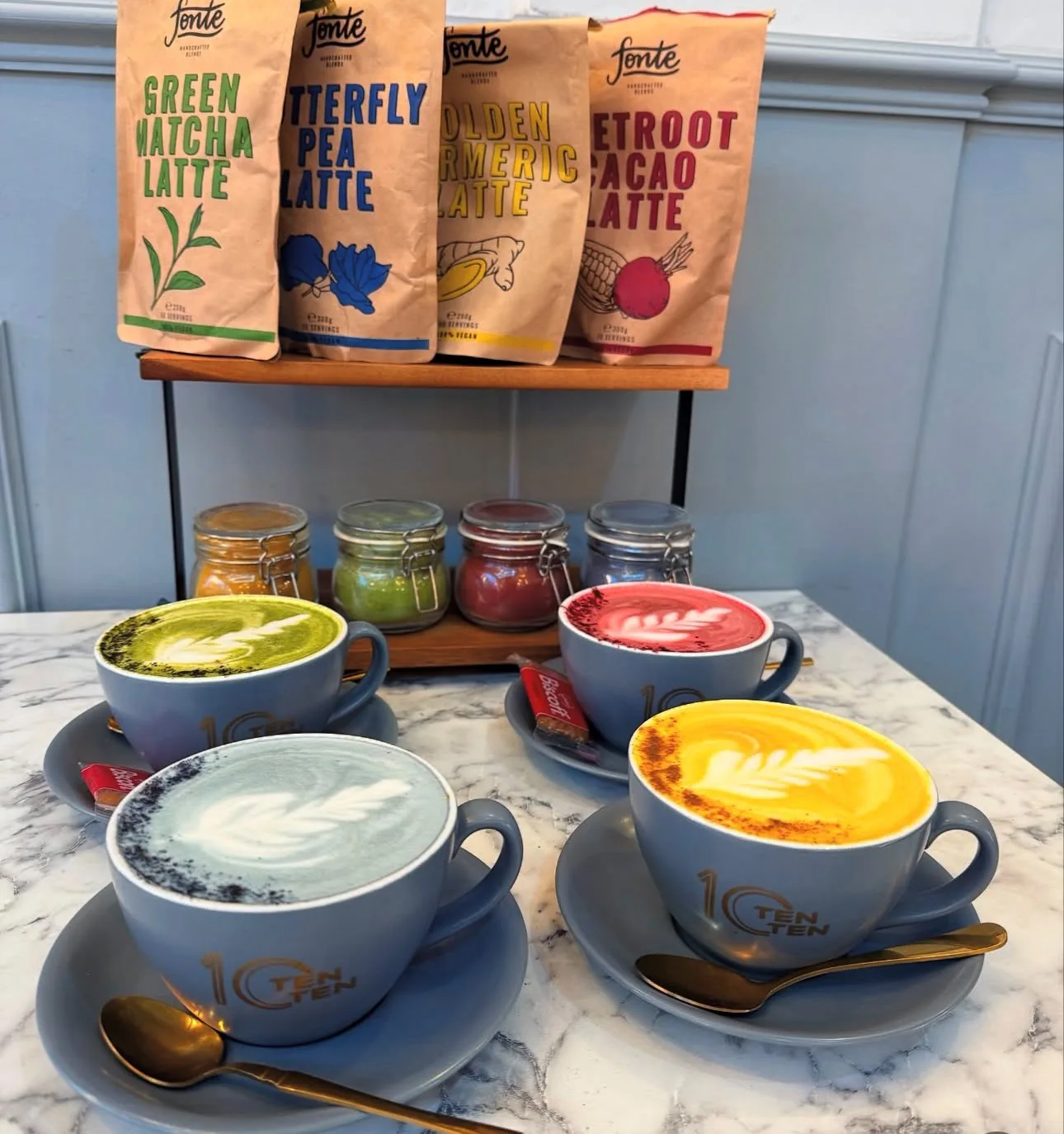 Four speciality lattes in TenTen's grey-blue branded cups — matcha green, butterfly-pea blue, beetroot pink and turmeric gold latte art — with jars of the powders on a stand behind.