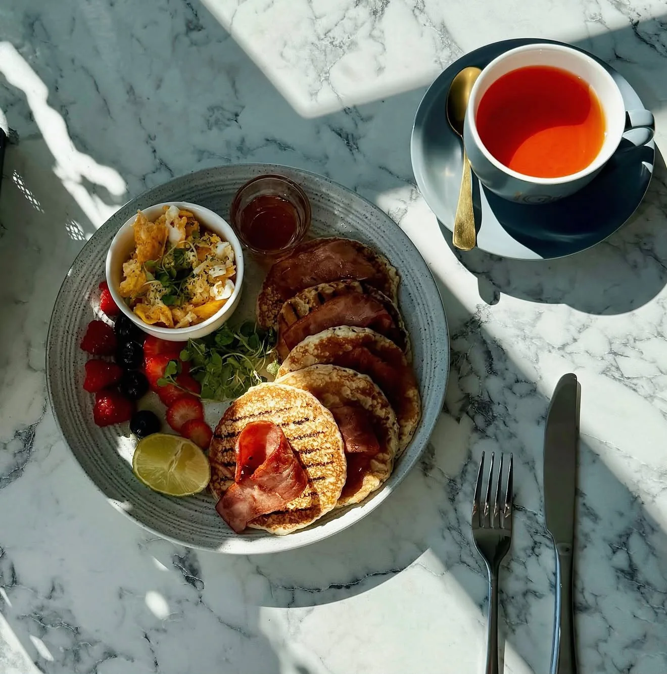Stack of grilled pancakes with turkey bacon, berries, scrambled eggs and a cup of tea in TenTen's grey-blue china
