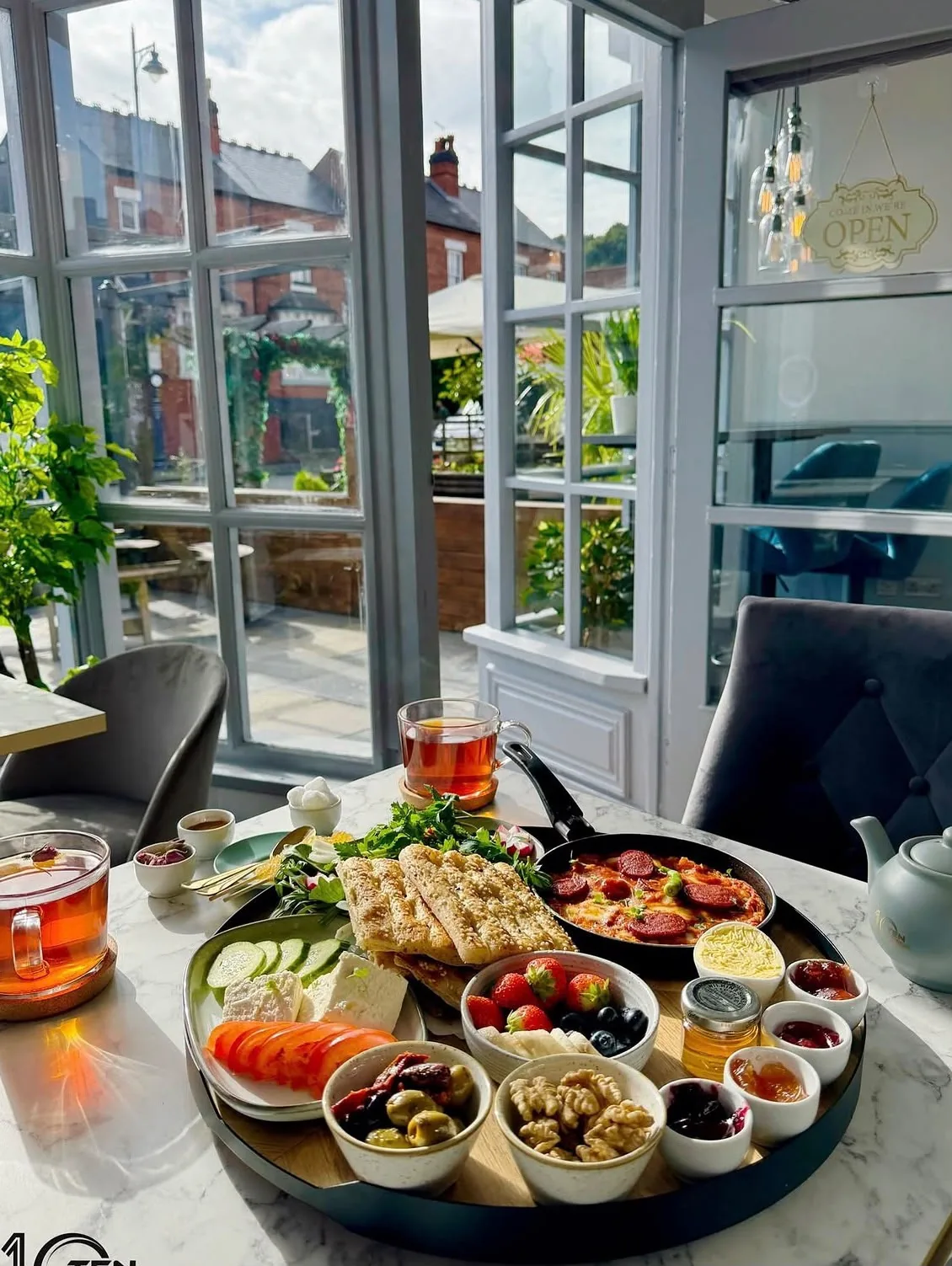 Persian sofre breakfast spread on a sunlit marble table at TenTen — flatbread, feta, honey pots and glasses of tea by the window, gold OPEN sign behind.