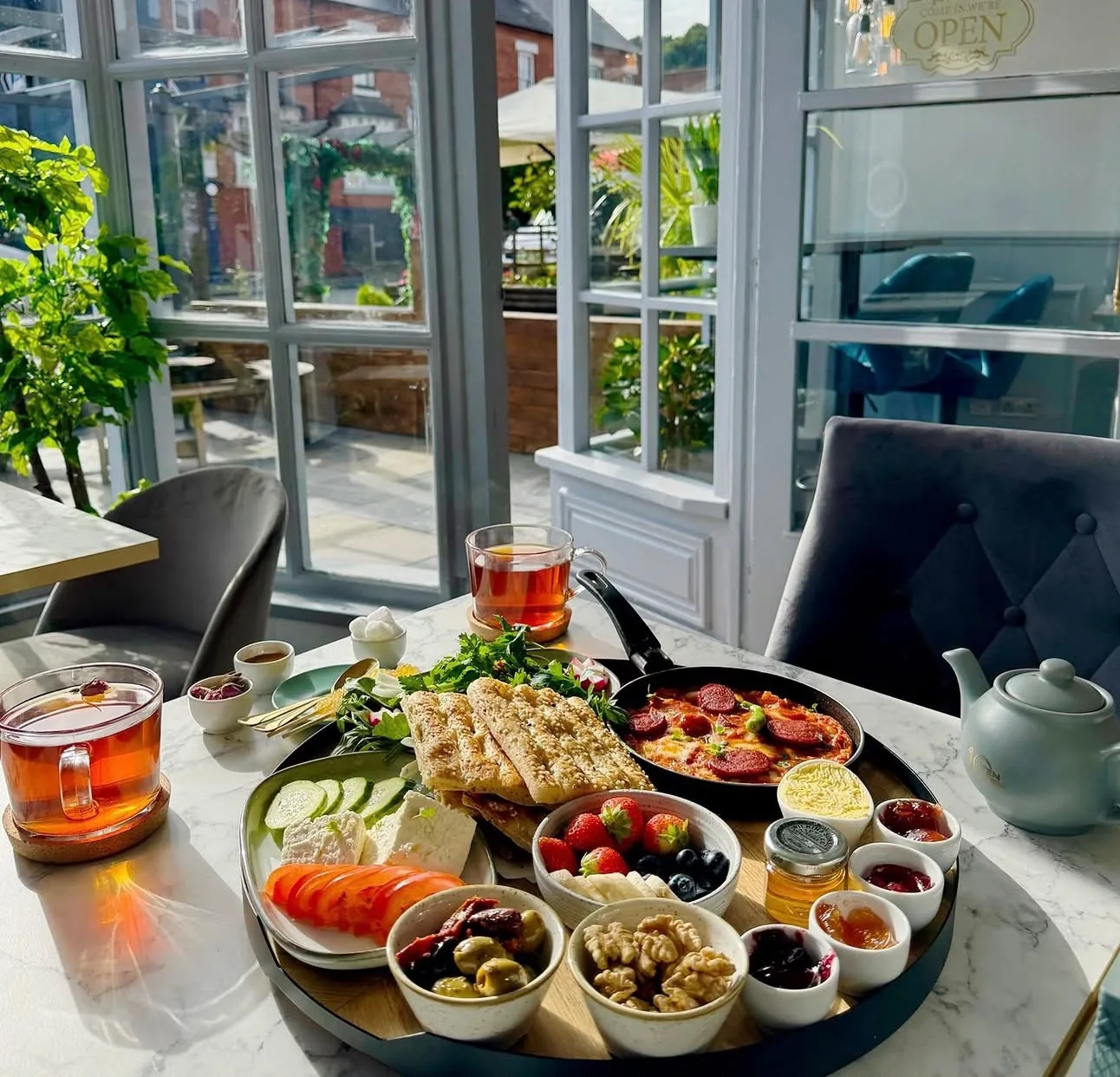 The full Persian sofre breakfast tray — sesame flatbread, feta, olives, walnuts, jams and a sausage-and-egg pan — with amber tea glasses on a marble table