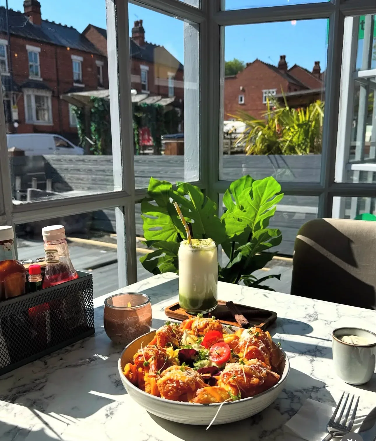 Sunny window corner at TenTen — loaded chips and an iced matcha on a marble table, a monstera plant and the red-brick terraces of Alcester Road beyond.